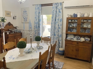 A dining area with a table set for four, surrounded by wooden chairs. A candelabra stands on the table, and a potted plant is on the floor. A wooden cabinet with glass doors displays various dishes.