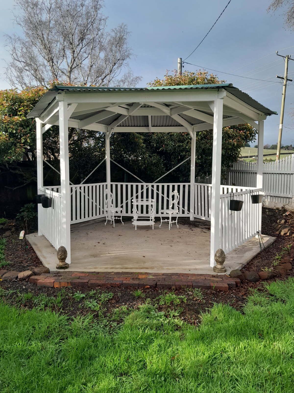 A covered patio with white railings and a roof, surrounded by a brick border and grass.