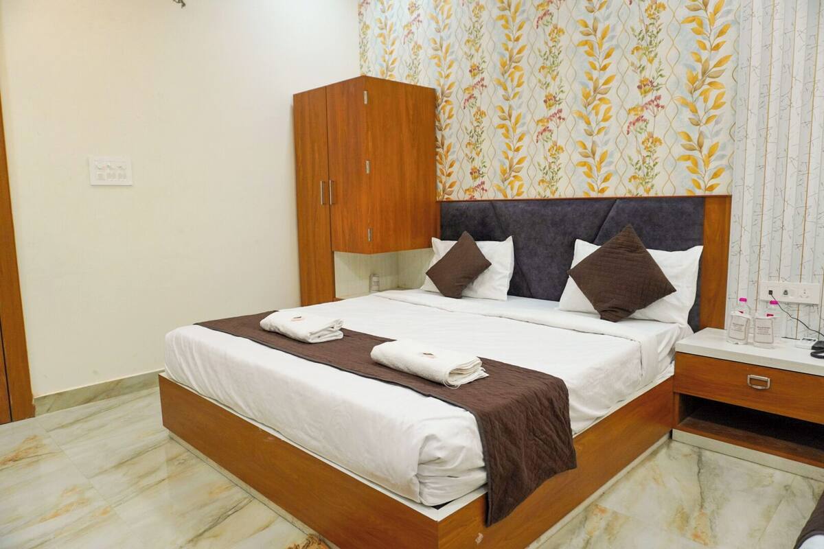 A neatly made bed with a brown blanket and two towels, a wooden wardrobe, and a bedside table with a bottle and a phone.