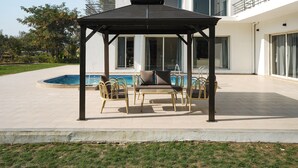 A covered outdoor seating area with a pool in the background. - Kings Villa (Bavla)