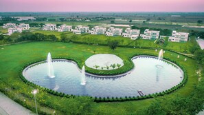 A large, circular water feature with fountains, surrounded by well-maintained lawns and a row of modern buildings in the background. - Kings Villa (Bavla)