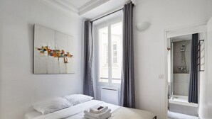 Hotellihuone, jossa on sänky, ikkuna verhoilla, suihkutila ja seinälle kiinnitetty taideteos.