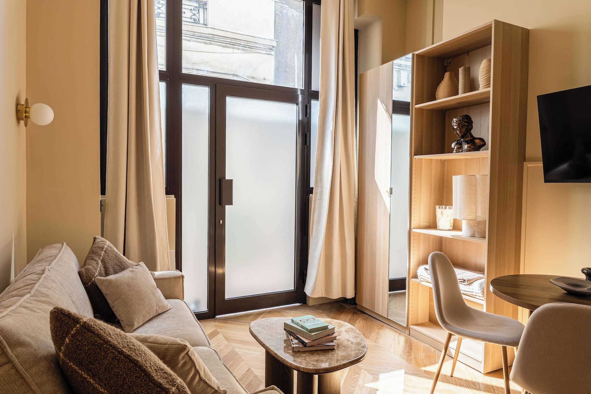 A modern living room with a sofa, a coffee table with books, a side table, a bookshelf, a television, and a sliding glass door leading to an outdoor area.