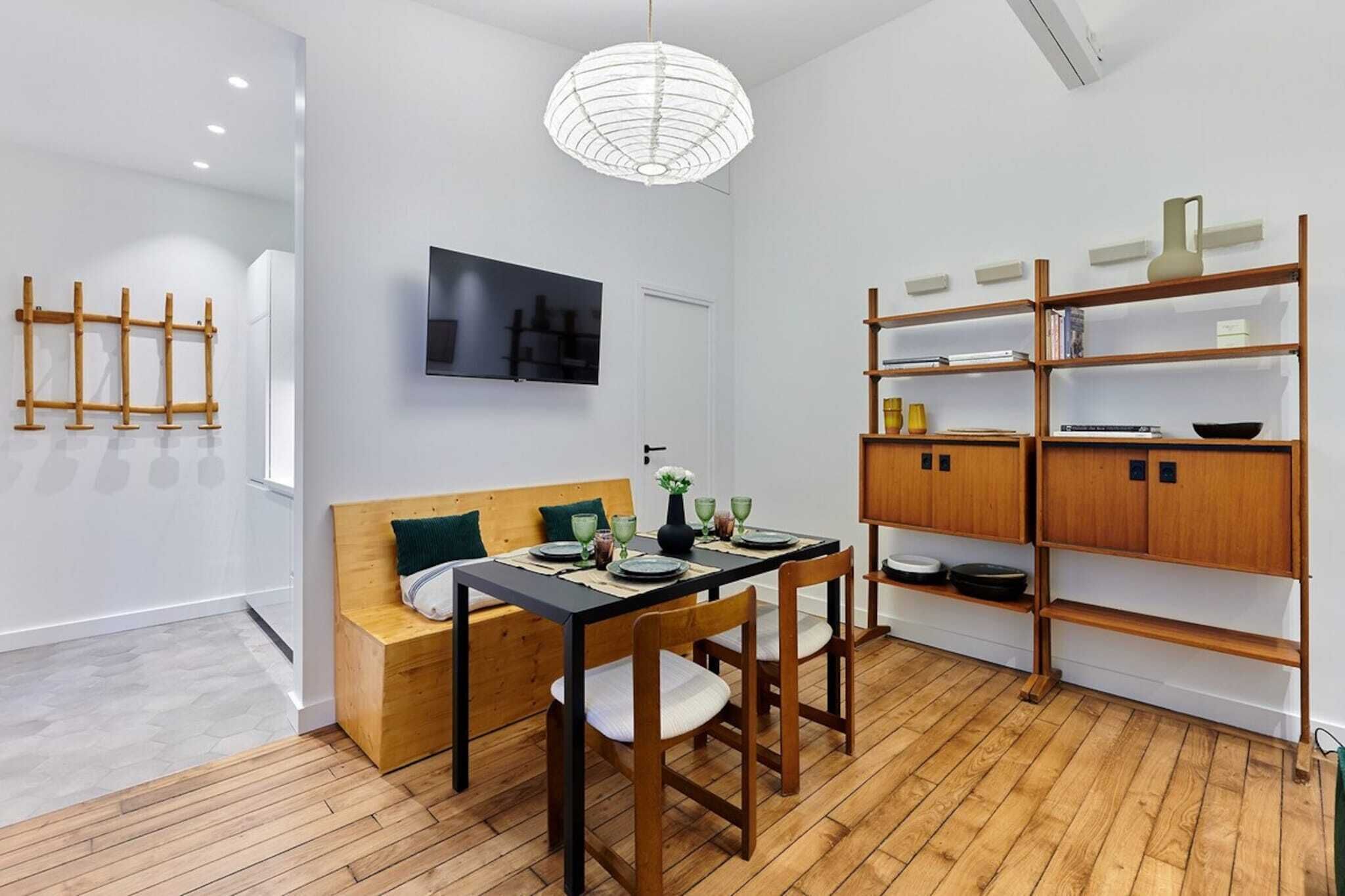 Un espace repas moderne avec une table en bois, des chaises et un banc. Un téléviseur est fixé au mur, et une bibliothèque en bois avec des étagères se trouve également dans la pièce.