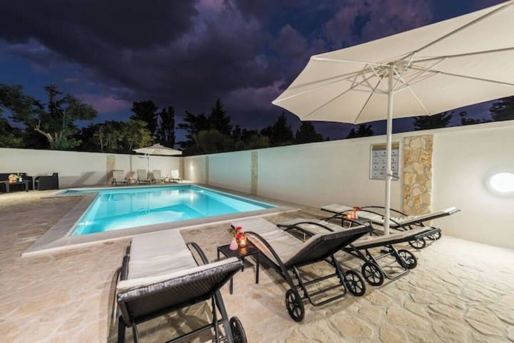 A swimming pool with lounge chairs and an umbrella, surrounded by a stone wall and trees.