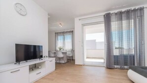 Una sala de estar moderna con televisor de pantalla plana, muebles blancos, un comedor con mesa y sillas, y un balcón con cortinas translúcidas.
