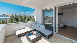 Un balcón con sofá y una mesa de centro, una puerta corrediza de vidrio que da a una habitación, y vistas a un edificio y árboles.