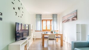 Une pièce avec un téléviseur posé sur un meuble blanc, une table à manger avec des chaises, un canapé bleu et une fenêtre avec des rideaux.