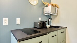 Cocina compacta con placa de inducción negra, un tostador y una cafetera. Hay dos enchufes y un estante pequeño con un objeto decorativo.