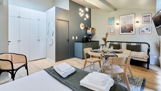 Un dormitorio moderno con cama, mesitas de noche, escritorio y silla. Cuenta con un armario, una pequeña kitchenette y un televisor montado en la pared.