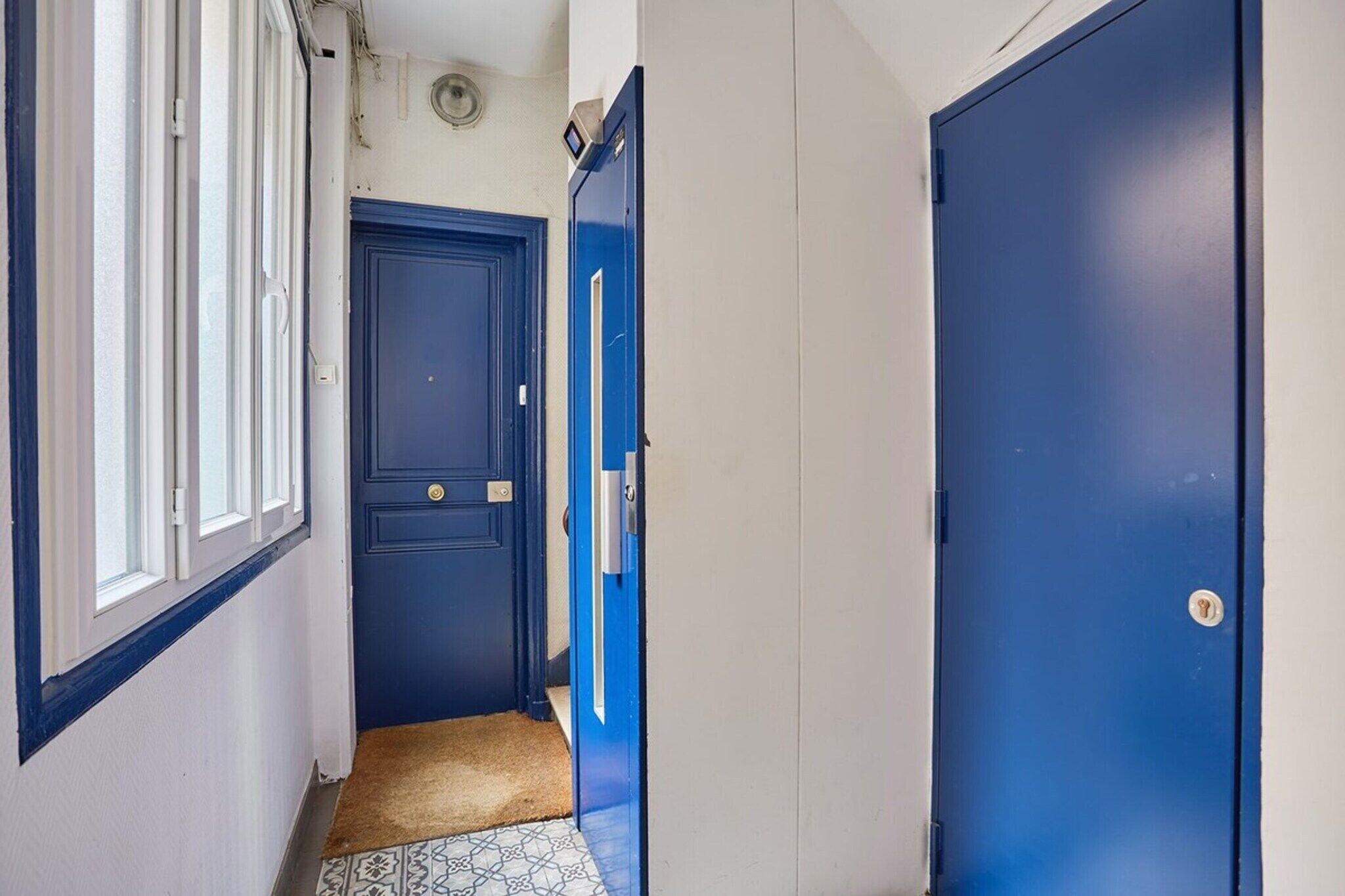 Ein schmaler Flur mit blauen Türen, einem Fenster und einer gemusterten Fußmatte.