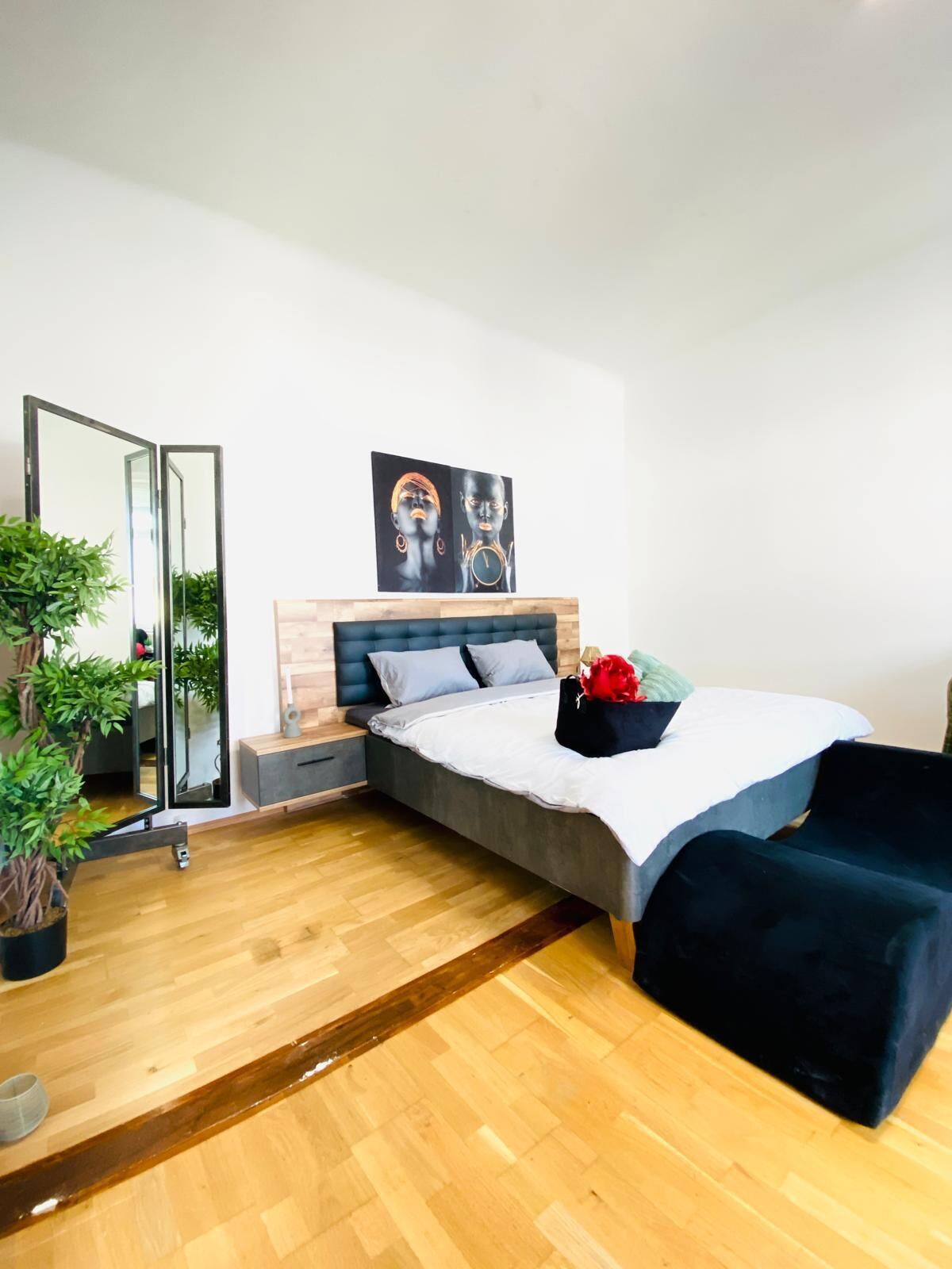 A modern bedroom with a bed, a bedside table, a mirror, a potted plant, and a painting on the wall.