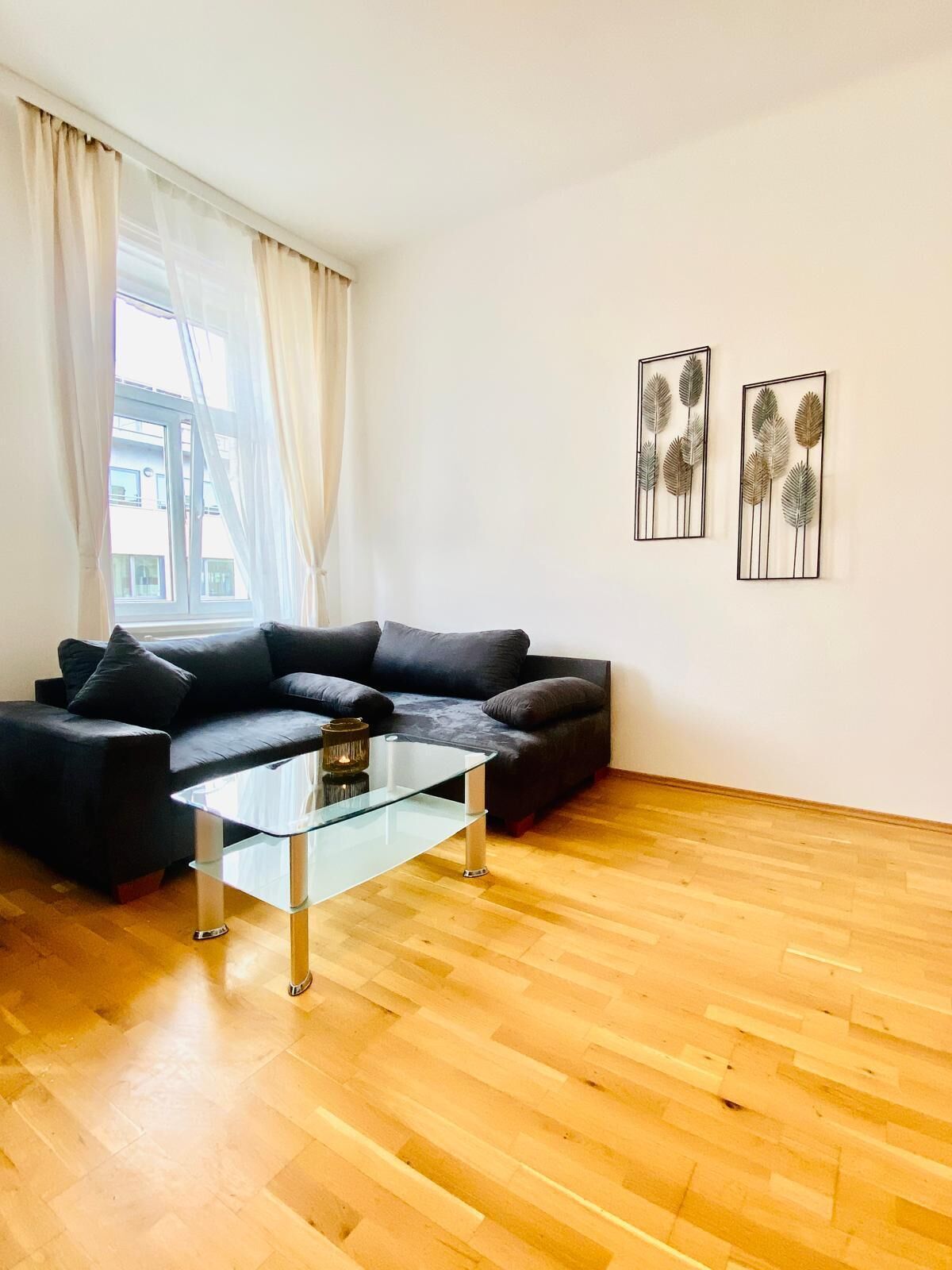 Una sala moderna con un sofá de cuero negro, una mesa de centro de vidrio y suelo de madera. Dos cuadros enmarcados decoran la pared sobre el sofá.