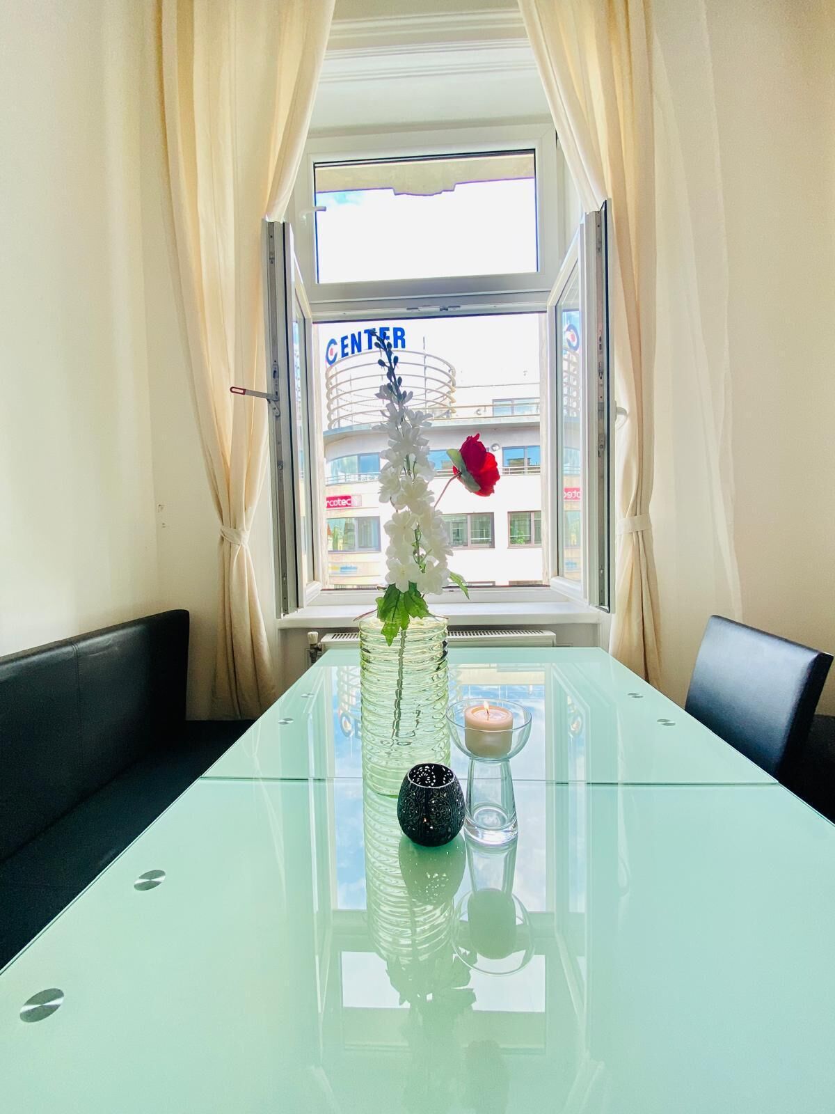 A glass table with a vase, candle, and decorative items. A red rose is on the table. The room has a window with curtains, a view of a building with a sign reading "CENTER".