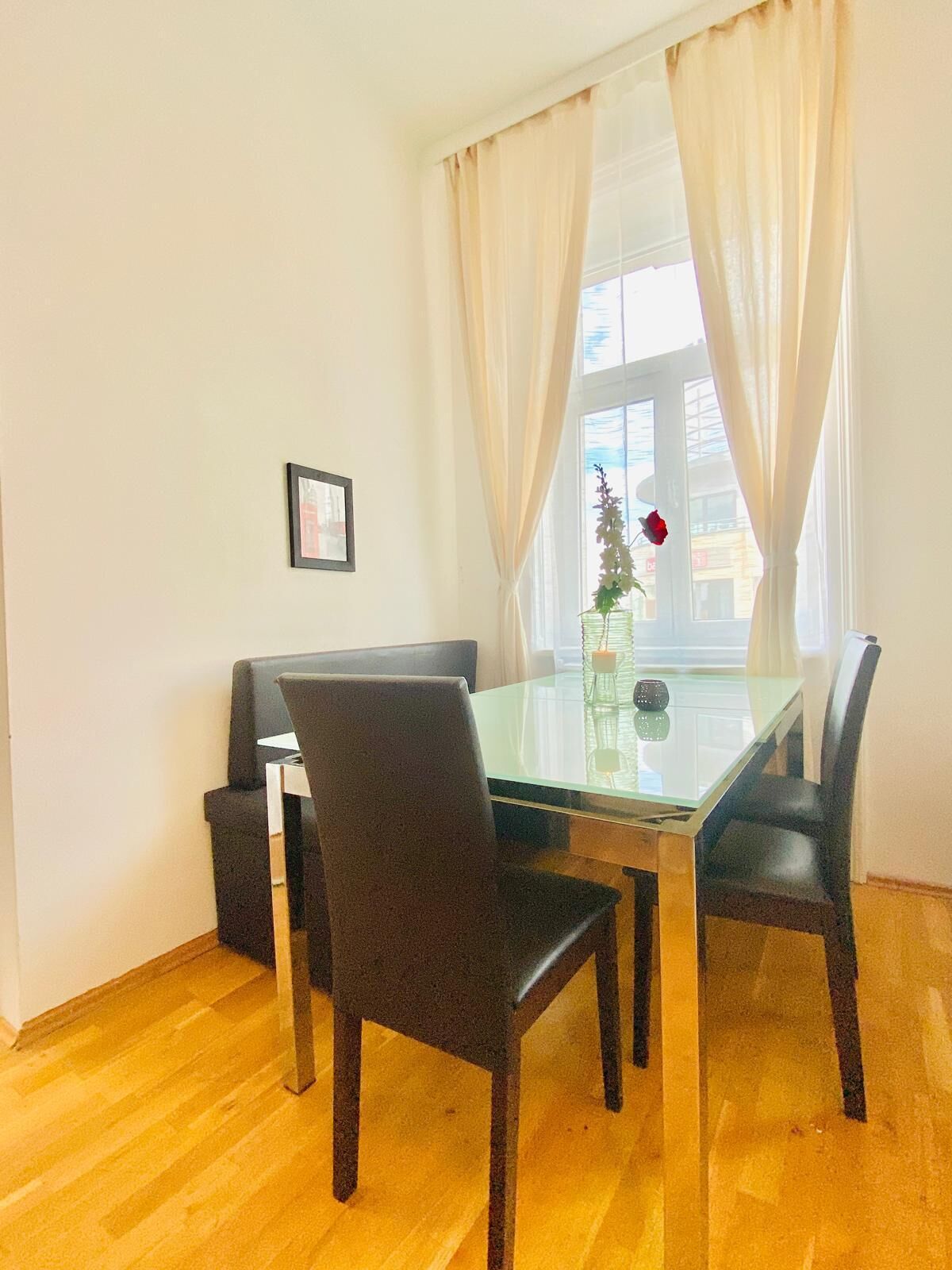 A dining area with a glass table and four chairs, a sofa, a framed picture on the wall, and a window with curtains.