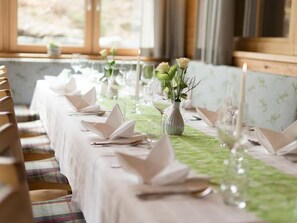 Una mesa de comedor decorada con servilletas blancas, cristalería y jarrones con flores, en una habitación con sillas de madera y una ventana.