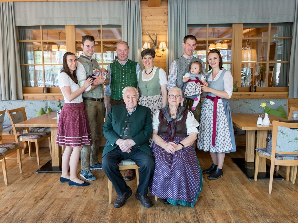 A family in traditional attire seated around a wooden table in a warmly lit room with large windows and wooden beams.