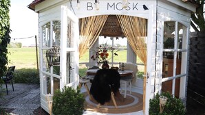 A cozy, rustic gazebo with a sign reading "BY MOSK," featuring a round table with chairs, a vase of flowers, and a birdcage. - Beautifully styled studio apartment (Betgummole)