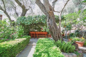 Um jardim com um banco e mesa de madeira, rodeado por vegetação exuberante e uma grande árvore.