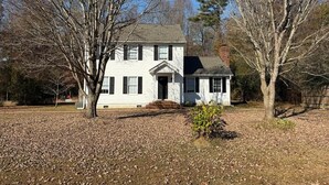 A white two-story house with a front porch, surrounded by leaf-covered ground and trees.
