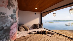 A bedroom with a large window overlooking a body of water, a bed with a floral-patterned headboard, and a view of boats and docks. - The Boat Company (Cherthala)