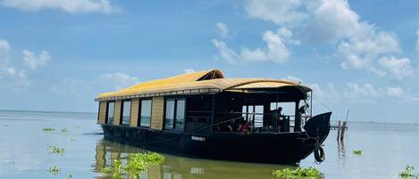 A boat with a covered deck and windows, floating on calm water with greenery around.