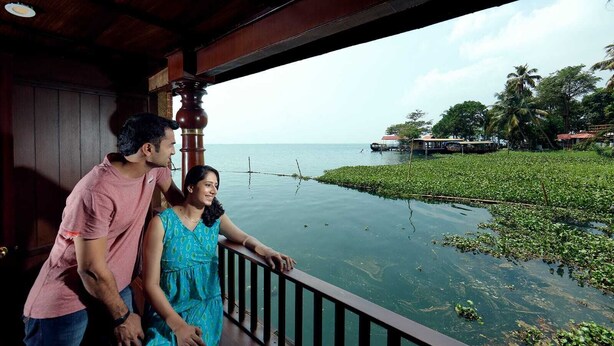 A wooden balcony overlooking a calm body of water with floating vegetation, a distant shoreline with buildings, and a clear sky. - The Boat Company (Cherthala)