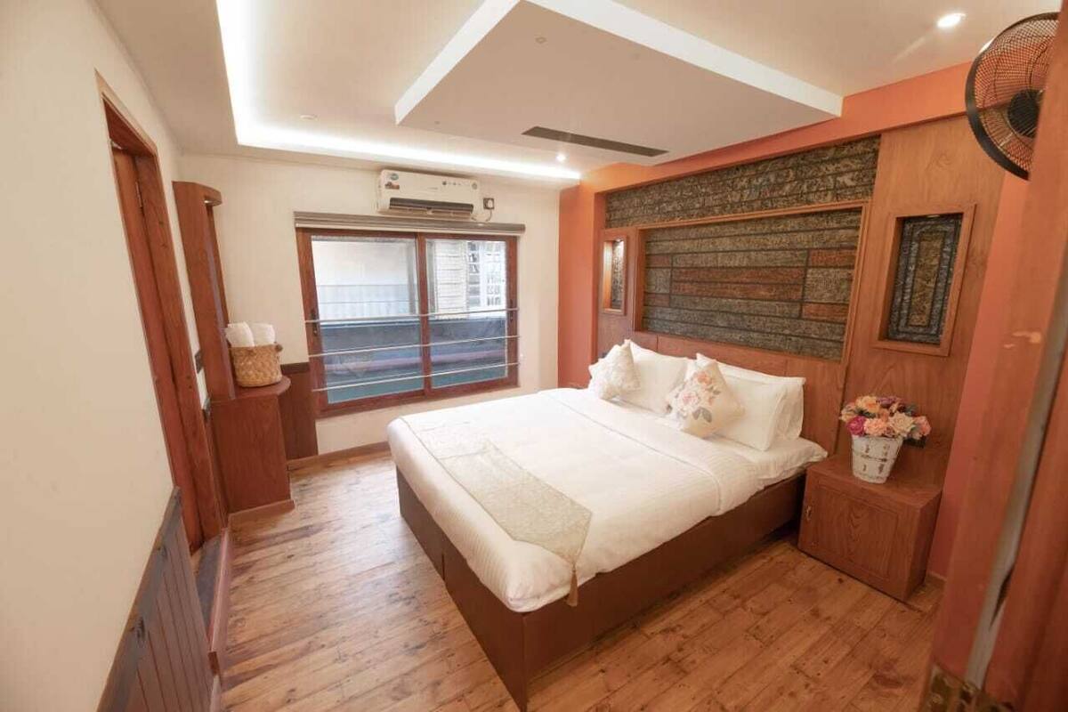 A bedroom with a large bed, wooden flooring, a window with blinds, and a wall-mounted air conditioner.