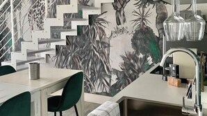Een moderne keuken met een trap waarop een tropische muurschildering is afgebeeld, een eethoek met groene stoelen, en een bar met een spoelbak en glazen.