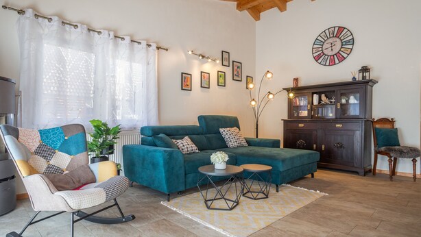 Ein Wohnzimmer mit einem türkisfarbenen Sofa, einem Schaukelstuhl, einem kleinen Tisch und einem Schrank mit einer Uhr an der Wand.