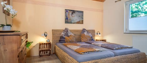 Una habitación con una cama grande de mimbre, cabecera de madera y un cuadro encima. Hay mesitas de noche con lámparas, un aparador y una ventana con persianas.