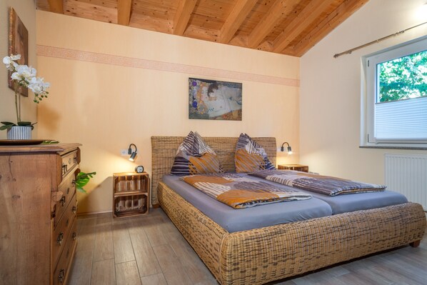 A room with a large wicker bed, wooden headboard, and a painting above it. There are bedside tables with lamps, a dresser, and a window with blinds.
