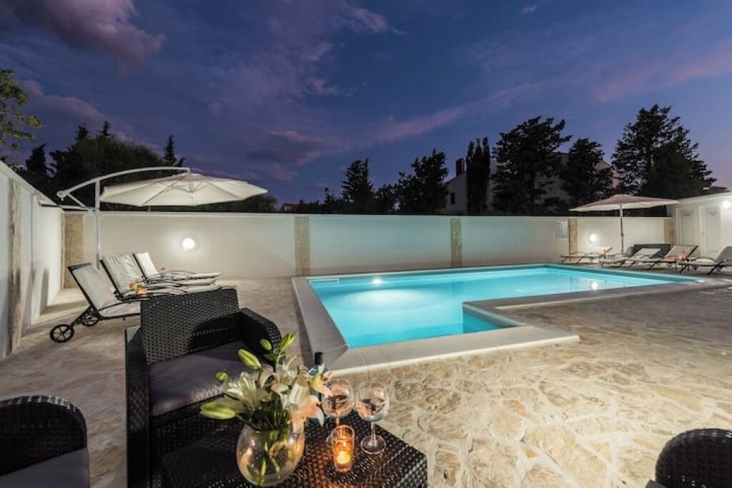 Une piscine entourée de chaises longues et de parasols, avec un sol en pierre et un ciel étoilé.