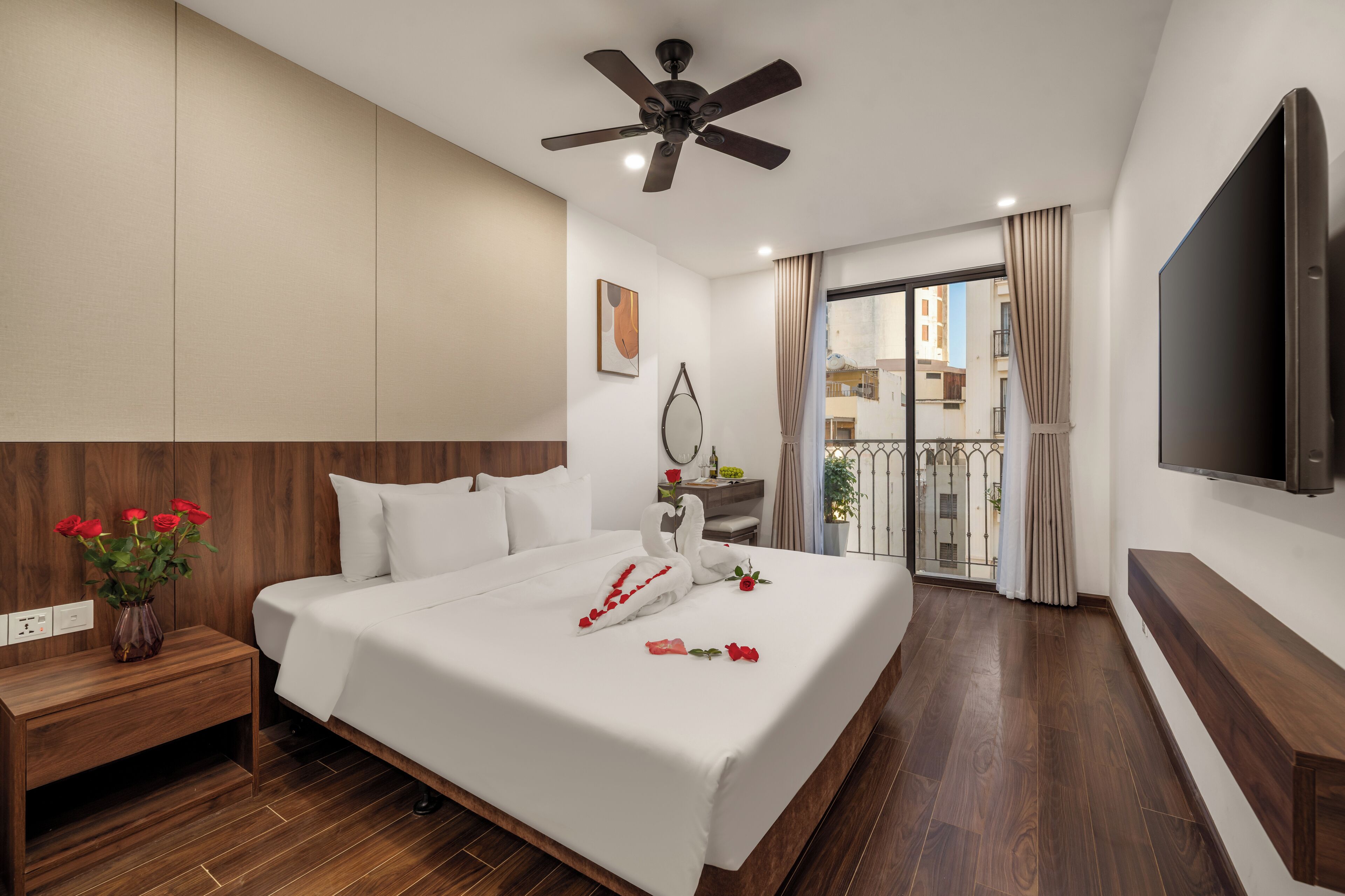 A hotel room with a bed, wooden headboard, a ceiling fan, a TV, a bedside table with a vase of flowers, and a view of the outside balcony.