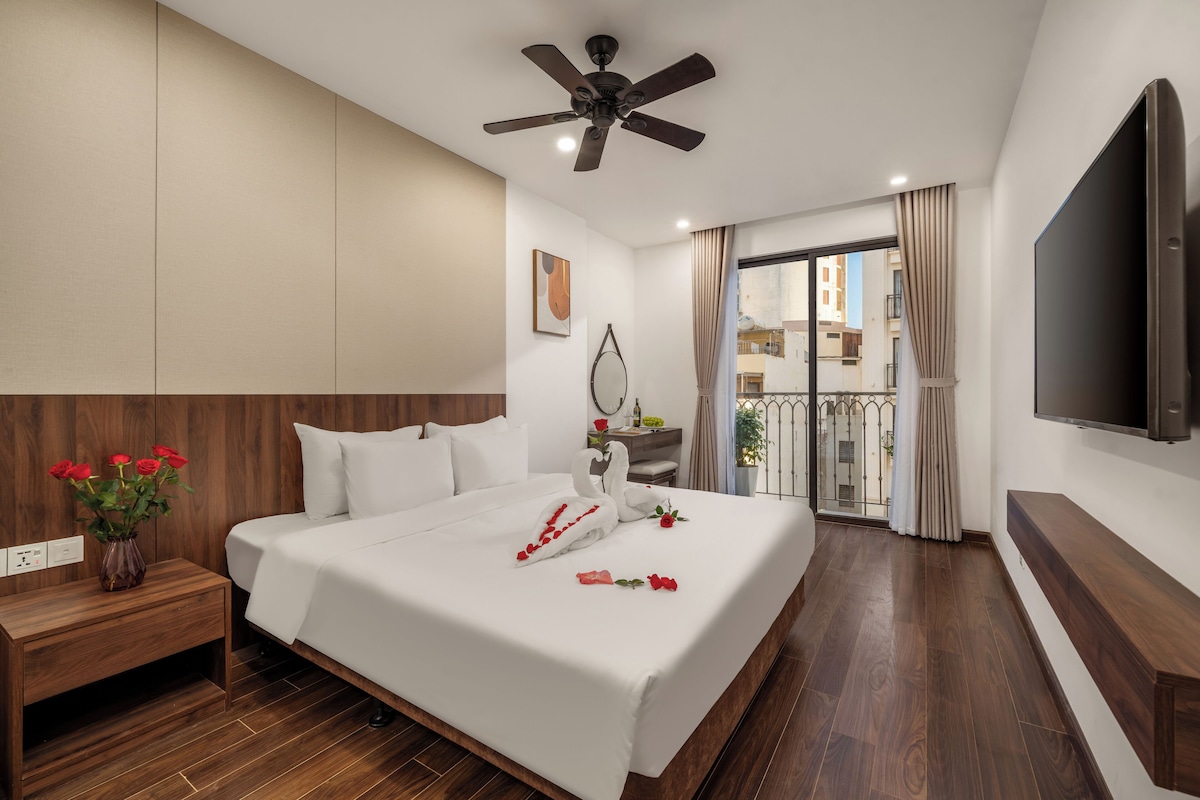 Camera d'albergo con un letto, una testiera in legno, una ventola a soffitto, un televisore, un comodino con un vaso di fiori e vista sul balcone esterno.