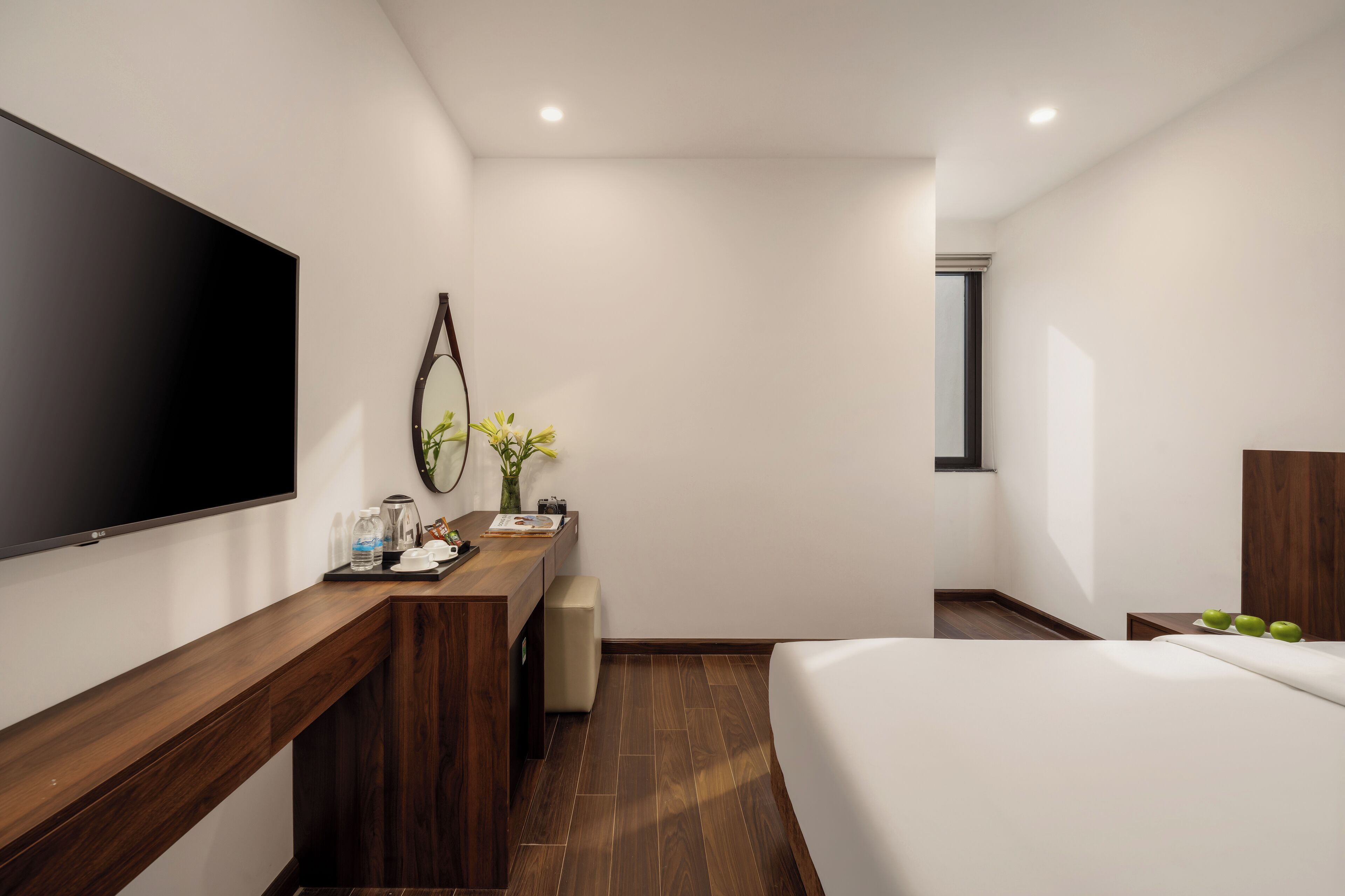 A modern hotel room with a large flat-screen TV, a wooden desk, a bedside table with apples, a bed, and a window.