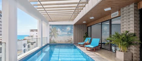 A rooftop infinity pool with a clear view of the ocean, lounge chairs, and a mural on the wall.