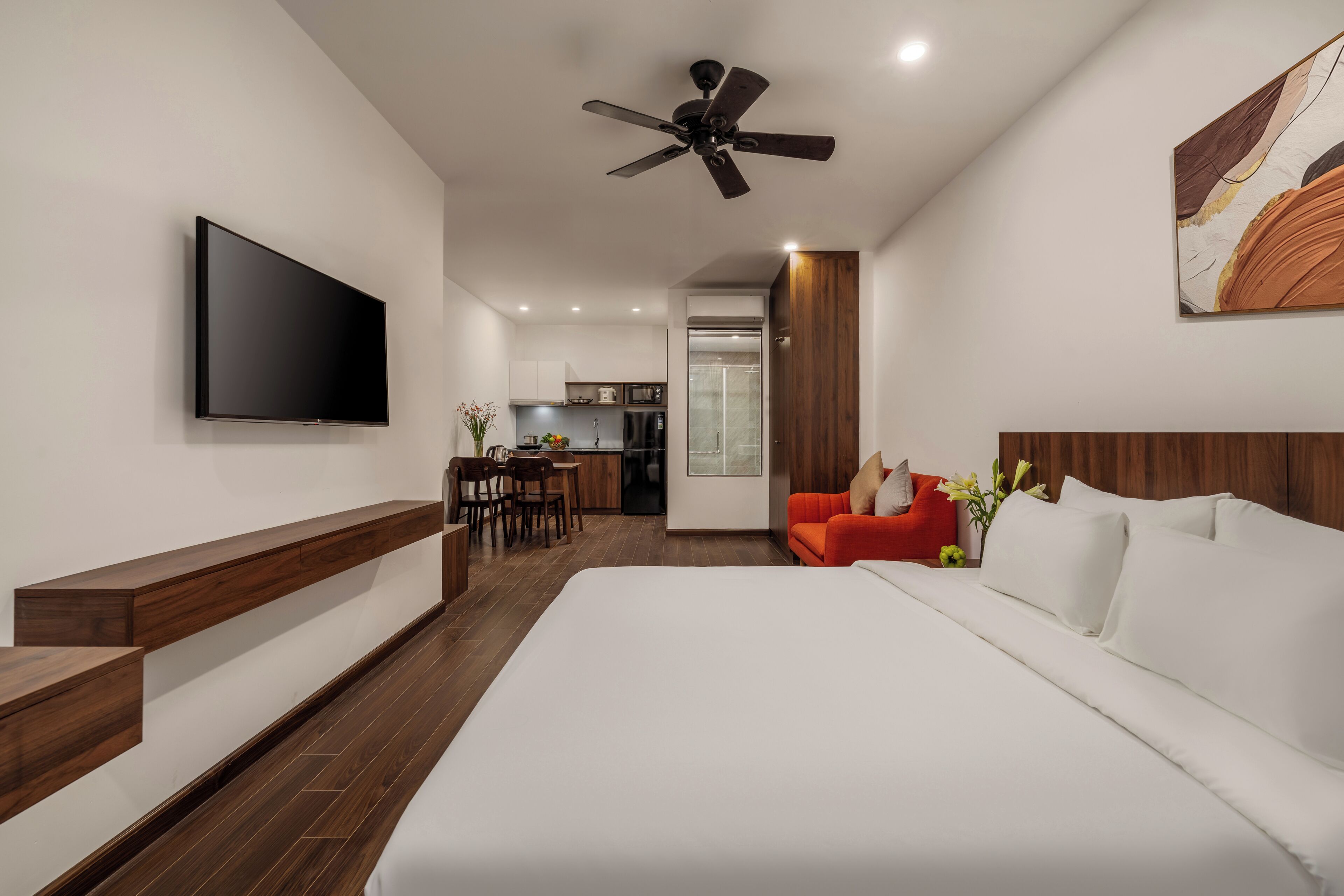 Una habitación de hotel moderna con una cama grande, un televisor de pantalla plana, un ventilador de techo, un banco de madera, un comedor y una cocina al fondo.