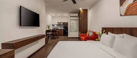Une chambre d’hôtel moderne avec un grand lit, un téléviseur à écran plat, un ventilateur de plafond, un banc en bois, un coin repas et une cuisine en arrière-plan.