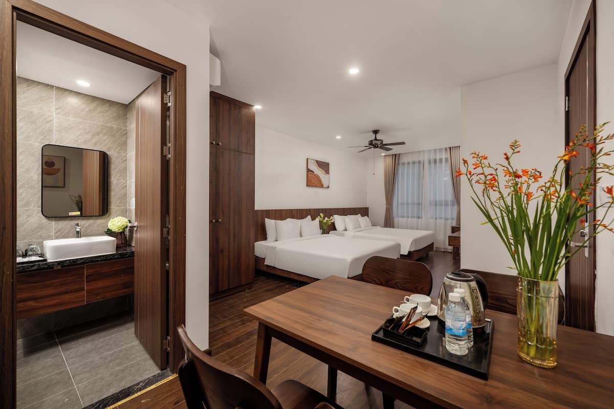 A hotel room with a wooden dining table, two beds, a bathroom with a sink and mirror, and a ceiling fan.