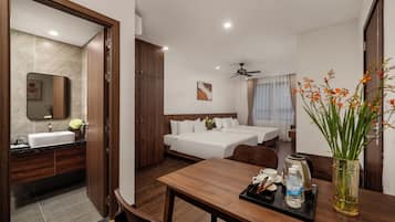 Una habitación de hotel con una mesa de comedor de madera, dos camas, un baño con lavamanos y espejo, y un ventilador de techo.