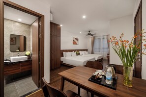 A hotel room with a wooden dining table, two beds, a bathroom with a sink and mirror, and a ceiling fan. - Zen Lotus Hotel & Apartment (Da Nang)