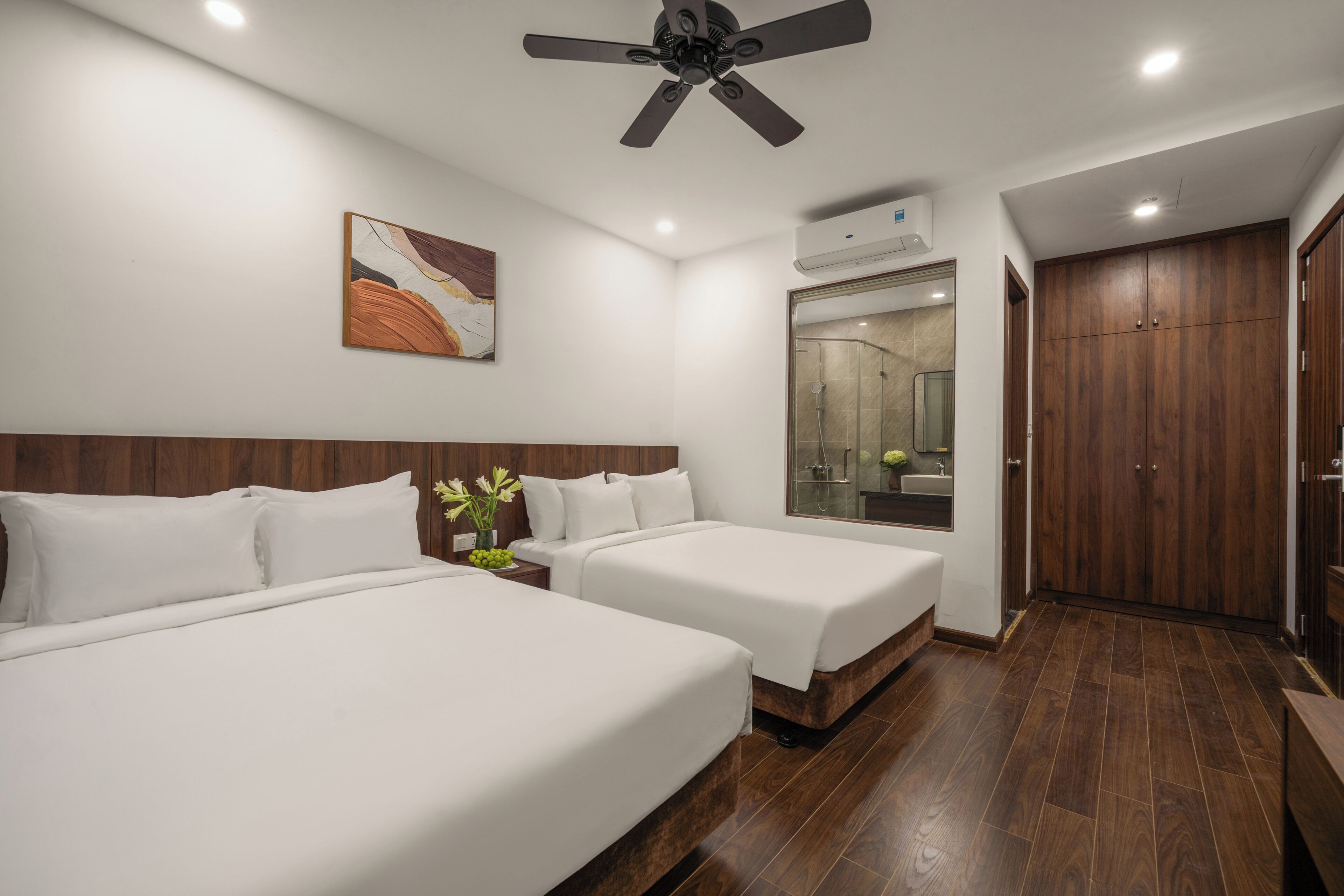 A hotel room with two beds, a ceiling fan, an air conditioning unit, a bathroom with a mirror, and wooden flooring.