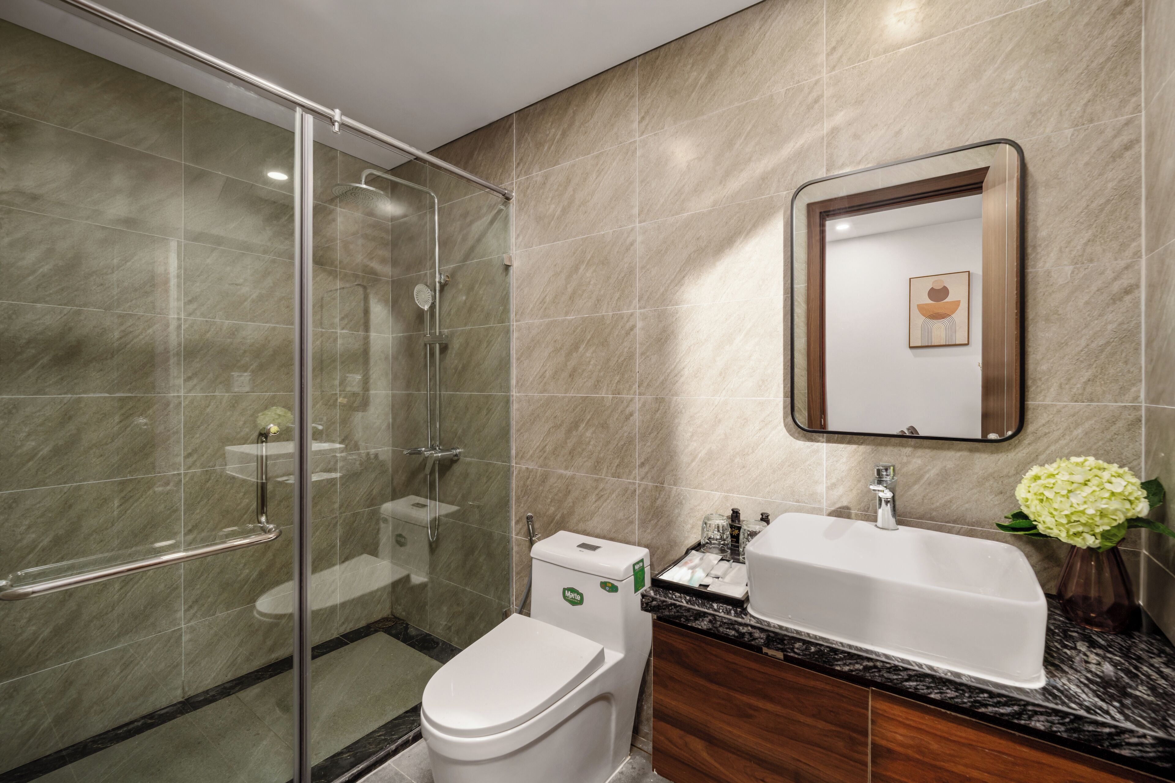 A modern bathroom with a glass-enclosed shower, a white toilet, a sink with a marble countertop, and a mirror with a framed picture.