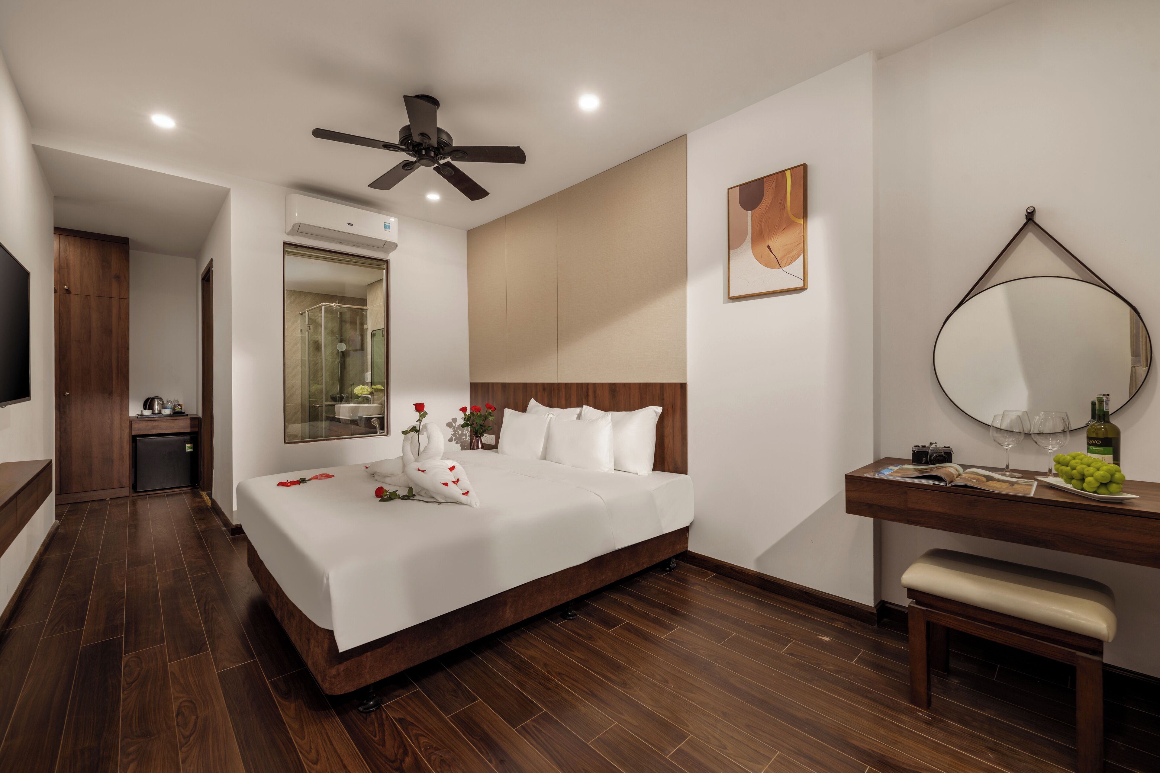 A modern hotel room with a large bed, a ceiling fan, a bathroom with a glass shower, a wooden bench, and a wooden vanity with a mirror.