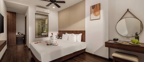 A modern hotel room with a large bed, a ceiling fan, a bathroom with a glass shower, a wooden bench, and a wooden vanity with a mirror.