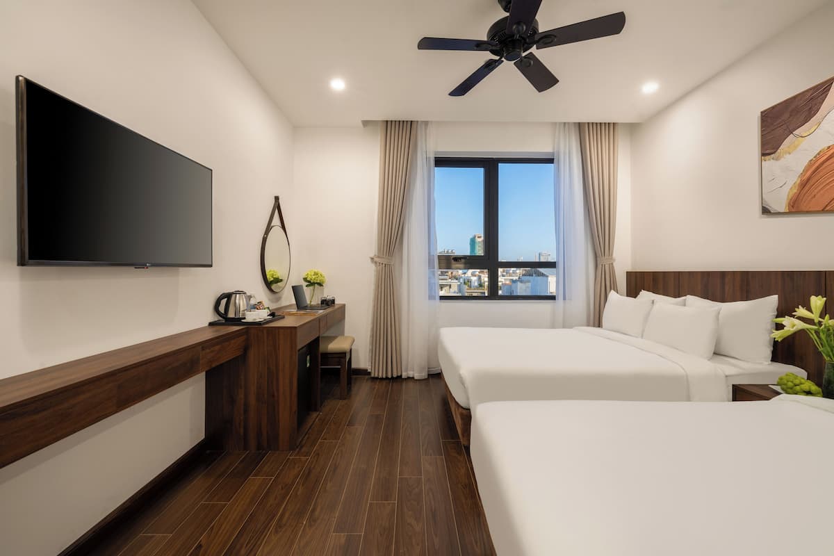 A hotel room with a flat-screen TV, a desk with a laptop, a chair, two beds, a ceiling fan, a window with curtains, and a city view.