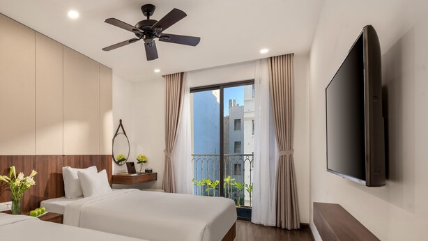 Ein Hotelzimmer mit zwei Betten, einem Deckenventilator, einem Fernseher, einem Balkon mit Vorhängen und Blick auf Gebäude.