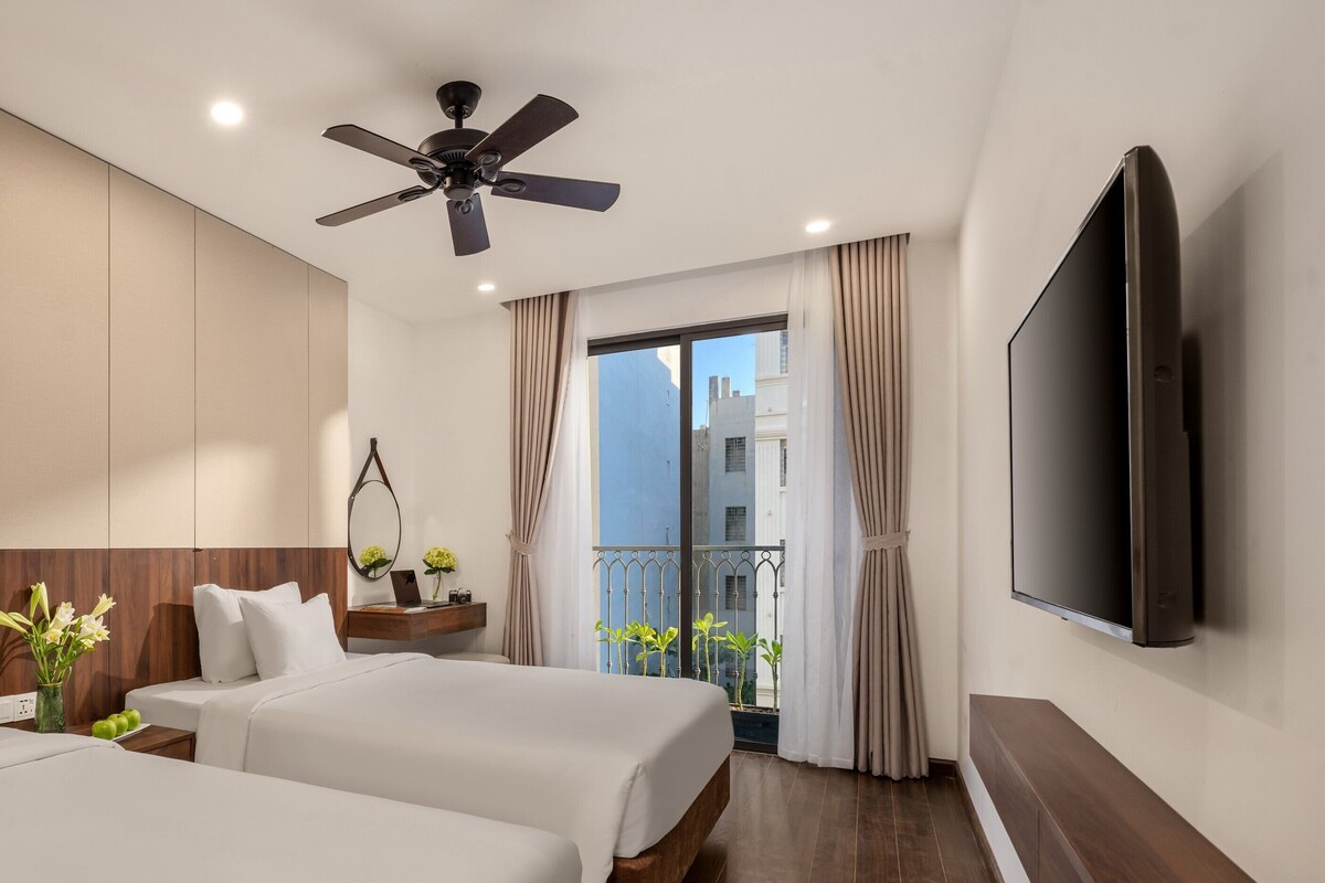 Habitación de hotel con dos camas, ventilador de techo, televisión, balcón con cortinas y vistas a edificios.