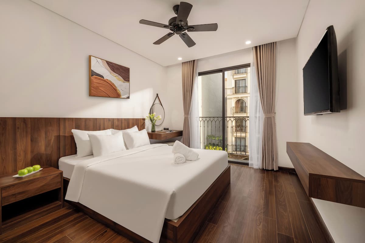 A hotel room with a large bed, a ceiling fan, a flat-screen TV, and a balcony with curtains.